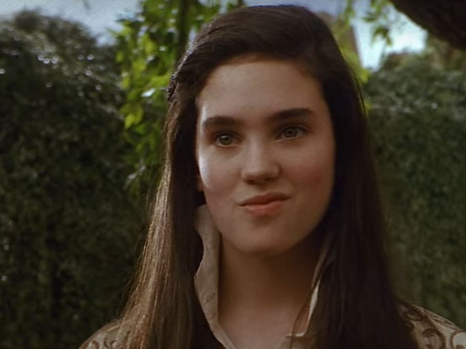 Image of Sarah in the 1986 film Labyrinth. She is a young girl with a determined smirk on her face, with a hedge maze in the background.